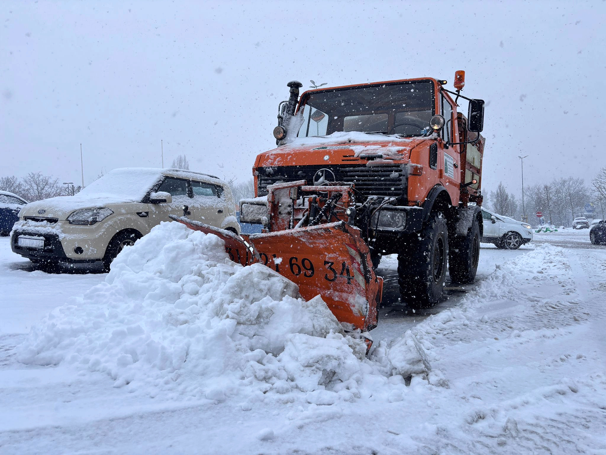 Orangefarbener Unimog der Baum- und Gartenpflege Kraus räumt bei starkem Schneefall einen Parkplatz.